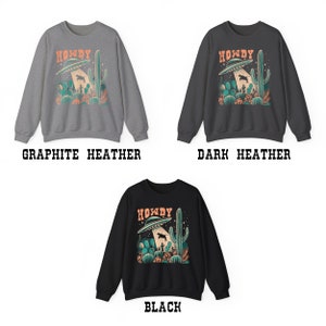 May include: A sweatshirt with a graphic print of a UFO over a desert landscape with the text "Howdy". The sweatshirt is available in three colors: graphite heather, dark heather, and black.