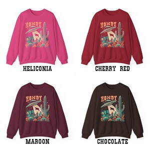 May include: Four different color sweatshirts with a graphic print of a UFO abducting a cow in a desert landscape with cacti. The text "Howdy" is printed above the UFO. The colors are Heliconia, Cherry Red, Maroon, and Chocolate.
