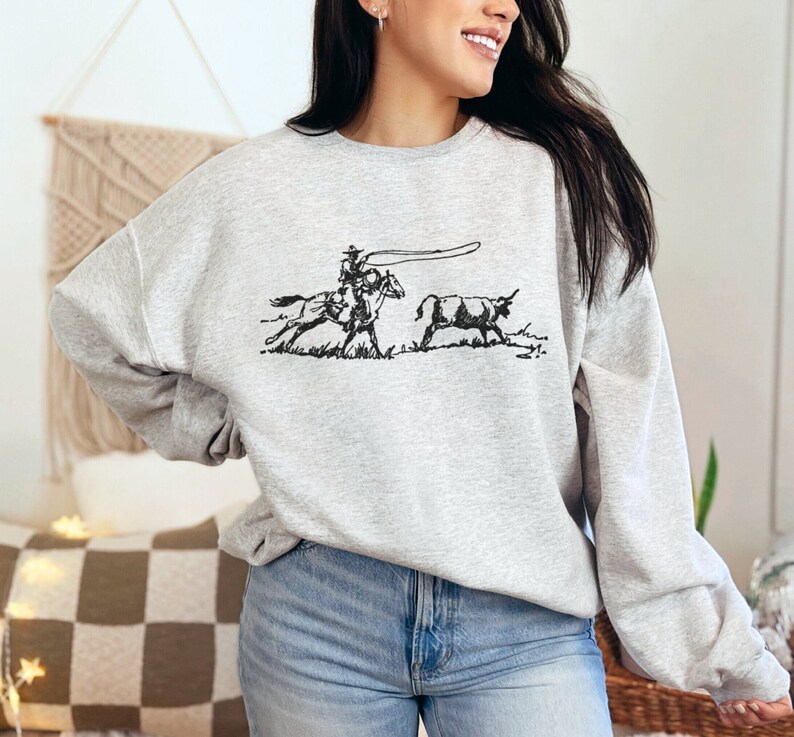 Peut inclure: Un sweat-shirt gris avec une illustration en noir et blanc d'un cow-boy &agrave; cheval qui l&acirc;che un lasso sur un taureau.