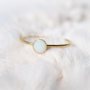 Op de afbeelding: Een delicate goudkleurige ring met een ronde, crèmekleurige, ondoorzichtige steen. De ring heeft een dunne, gepolijste band, met de steen in een eenvoudig, elegant ontwerp gezet. De ring wordt getoond op een zacht, wit, getextureerd oppervlak.