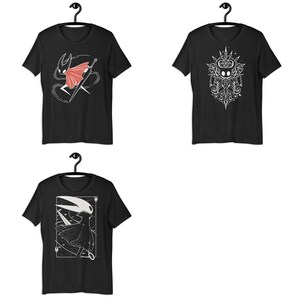 May include: Three black short-sleeve t-shirts displayed on hangers. The top left shirt features a red and white character graphic. The top right shirt shows a white intricate symmetrical design. The bottom shirt displays a white character illustration.