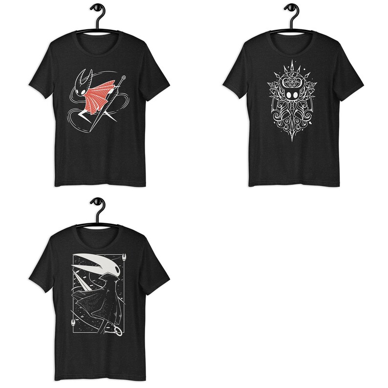 May include: Three black short-sleeve t-shirts displayed on hangers. The top left shirt features a red and white character graphic. The top right shirt shows a white intricate symmetrical design. The bottom shirt displays a white character illustration.