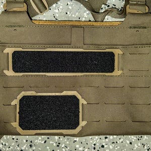 May include: Tan chest rig with two black hook-and-loop panels and tan accents. The rig has adjustable straps and buckles. The design includes multiple rows of horizontal slots for attaching additional gear. The rig is designed for tactical use.
