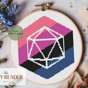 Op de afbeelding: Een kruissteekpatroon in een houten hoepel heeft een geometrisch ontwerp met roze, paarse, zwarte en blauwe secties. Een witte, geometrische vorm staat in het midden. De tekst "PDF PATTERN" en "the COZY READER SOCIETY" zijn ook zichtbaar.