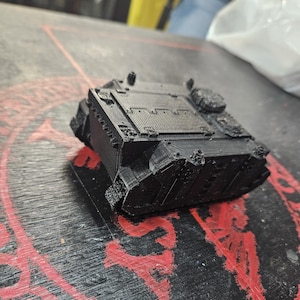 Puede incluir: Un modelo de tanque en miniatura impreso en 3D de color negro, con un chasis rectangular, paneles superiores texturizados, blindaje lateral y detalles de escotillas circulares. El objeto muestra l&iacute;neas de capa del proceso de impresi&oacute;n.
