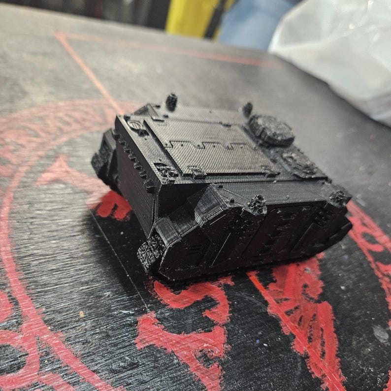 Puede incluir: Un modelo de tanque en miniatura impreso en 3D de color negro, con un chasis rectangular, paneles superiores texturizados, blindaje lateral y detalles de escotillas circulares. El objeto muestra l&iacute;neas de capa del proceso de impresi&oacute;n.