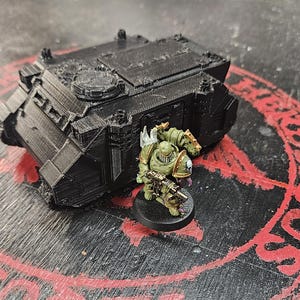 Puede incluir: Un veh&iacute;culo de transporte blindado en miniatura impreso en 3D de color negro con una superficie texturizada, colocado junto a una peque&ntilde;a figura de soldado pintada de verde que sostiene un rifle y est&aacute; sobre una base circular negra.