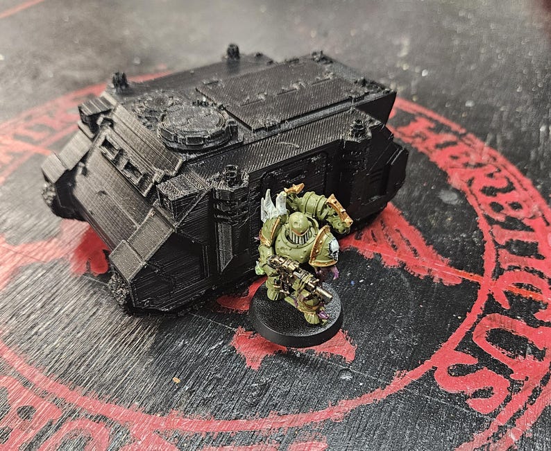 Puede incluir: Un veh&iacute;culo de transporte blindado en miniatura impreso en 3D de color negro con una superficie texturizada, colocado junto a una peque&ntilde;a figura de soldado pintada de verde que sostiene un rifle y est&aacute; sobre una base circular negra.