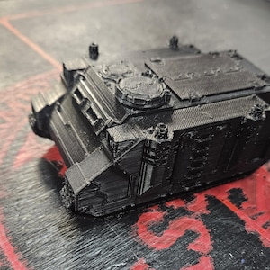 Puede incluir: Un veh&iacute;culo de transporte blindado en miniatura impreso en 3D de color negro, con un chasis cuadrado, una cubierta superior plana con detalles de escotillas circulares, paneles laterales y una placa de blindaje frontal inclinada con ranuras.