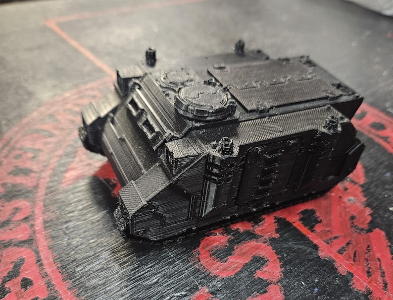 Puede incluir: Un veh&iacute;culo de transporte blindado en miniatura impreso en 3D de color negro, con un chasis cuadrado, una cubierta superior plana con detalles de escotillas circulares, paneles laterales y una placa de blindaje frontal inclinada con ranuras.