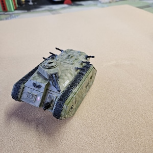 May include: A miniature military tank toy in shades of green and tan, with black detailing. The tank has multiple turrets and a textured surface, with the word "CADIA" printed on the side. The tank is set against a neutral background.
