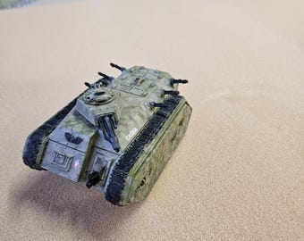 May include: A miniature military tank toy in shades of green and tan, with black detailing. The tank has multiple turrets and a textured surface, with the word "CADIA" printed on the side. The tank is set against a neutral background.