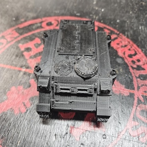 Puede incluir: Un veh&iacute;culo blindado en miniatura impreso en 3D de color negro, con un chasis rectangular, una escotilla superior plana y dos puertos de torreta circulares. La superficie muestra l&iacute;neas de capa visibles del proceso de impresi&oacute;n y textura.