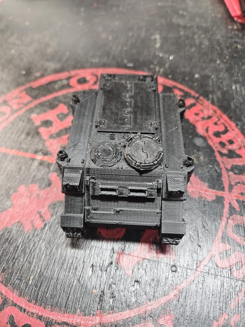 Puede incluir: Un veh&iacute;culo blindado en miniatura impreso en 3D de color negro, con un chasis rectangular, una escotilla superior plana y dos puertos de torreta circulares. La superficie muestra l&iacute;neas de capa visibles del proceso de impresi&oacute;n y textura.