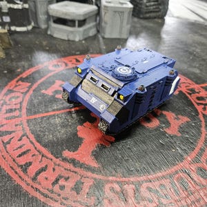 Puede incluir: Un veh&iacute;culo de transporte blindado en miniatura de color azul con el n&uacute;mero romano IV en la parte delantera, faros amarillos y una escotilla circular en la parte superior. El modelo tiene paneles marrones texturizados y detalles met&aacute;licos.