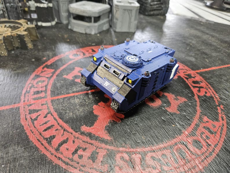 Puede incluir: Un veh&iacute;culo de transporte blindado en miniatura de color azul con el n&uacute;mero romano IV en la parte delantera, faros amarillos y una escotilla circular en la parte superior. El modelo tiene paneles marrones texturizados y detalles met&aacute;licos.