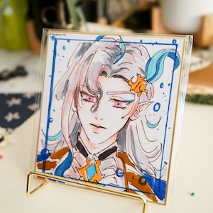 以下が含まれることがあります： アニメスタイルのキャラクターイラストが描かれた正方形のアートプリント。キャラクターは銀色の髪、ピンクの目、青とオレンジのアクセントが特徴です。アートワークは透明な保護スリーブと金色の金属製スタンドに展示されています。