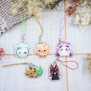 May include: Five colorful acrylic charms with cartoon animal designs, each attached to a lanyard. The charms feature a light blue bunny, an orange bird, a purple creature, a brown bunny, and a black cat. The charms are on a white surface with floral accents.