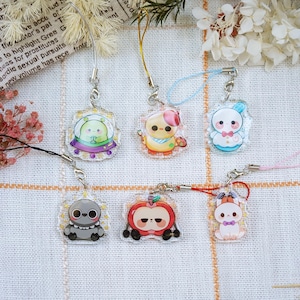 May include: A collection of six acrylic charms with cartoon character designs. The charms feature various creatures, including an alien in a UFO, a chick with a pink bow, and a bunny. Each charm has a different colored lanyard.