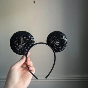 May include: A black sequin mouse ear headband. The ears are round and covered in shiny black sequins. The headband is black and has a simple, curved design. Ideal for themed events or adding a touch of fun to any outfit.