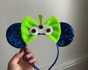 Puede incluir: Una diadema azul con orejas de lentejuelas y un lazo verde neón. El lazo presenta una cara alienígena en blanco y negro con ojos grandes y una antena verde. Las orejas son de color azul oscuro.