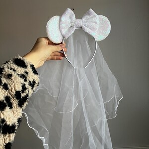 May include: A white sequined headband with large mouse ears and a bow, paired with a sheer, layered veil. The headband is silver-toned, and the veil is a soft, flowing fabric. This bridal accessory is perfect for a themed wedding or hen party.