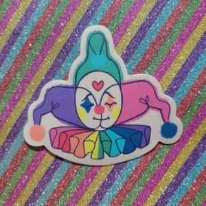 May include: A colourful sticker of a jester with a rainbow ruff, blue hat, and a heart on its nose. The jester has a purple and pink jester hat with a blue pom-pom.