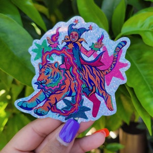 Puede incluir: Una pegatina holográfica con un bufón montando un tigre. El bufón lleva un sombrero azul y rosa y una capa roja y rosa. El tigre es naranja y negro con rayas azules y rosas. La pegatina tiene un acabado brillante.
