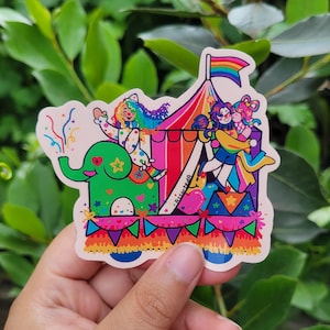 Può includere: Un adesivo colorato raffigurante una scena circense con una bandiera arcobaleno. L'adesivo raffigura un elefante verde, clown e un tendone da circo. Il design include cuori, stelle e coriandoli, con un'estetica vivace e giocosa.