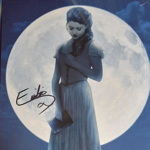 May include: A woman in a long, flowing white dress stands in front of a large, bright moon. She is holding a book in her hand and her head is bowed. The background is a dark blue sky with a hint of water at the bottom of the image. The woman's signature is visible in the lower left corner of the image.