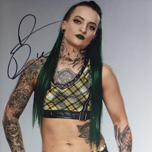 May include: A woman with green hair and tattoos wearing a yellow and black plaid crop top, a black belt with silver buckles, and black and yellow plaid shorts. She is posing with her hands on her hips.