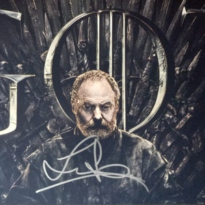 May include: A black and white photo of a man with a beard sitting on a throne made of swords. The throne is shaped like a chair with a large back and armrests. The man is wearing a dark jacket and has a serious expression on his face. The photo is signed with the name "Peter Dinklage".
