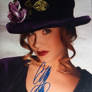 Puede incluir: Una mujer con cabello rubio que lleva un sombrero de terciopelo morado con un diseño de hojas doradas y una flor morada. Lleva una blusa morada y tiene una firma azul en el pecho.