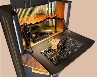 May include: A black and gold bar cabinet with a fold-down countertop. The cabinet has a wood interior with shelves and a built-in bottle opener. The countertop has a black and gold geometric pattern. A cocktail glass with a cherry garnish sits on the countertop.
