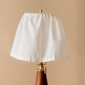 May include: A table lamp with a white fabric lampshade. The lamp has a wooden base and brass accents. The lampshade is a simple, cone-shaped design. The lamp is turned on, and the light is shining through the shade.