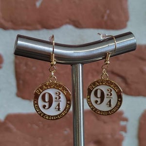 Peut inclure: Boucles d'oreilles dorées avec un charme en émail blanc portant le texte "Platform 9 3/4 King's Cross London".