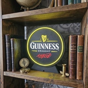 Peut inclure: Un panneau Guinness Draught avec une bordure n&eacute;on jaune est pr&eacute;sent&eacute; sur une &eacute;tag&egrave;re en bois. Le panneau est noir avec des lettres blanches et un logo de harpe dor&eacute;e. L'&eacute;tag&egrave;re contient &eacute;galement des livres anciens, une boussole et un globe.