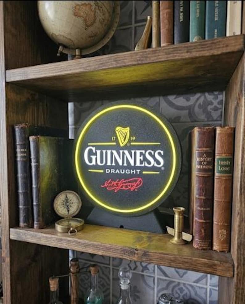 Peut inclure: Un panneau Guinness Draught avec une bordure n&eacute;on jaune est pr&eacute;sent&eacute; sur une &eacute;tag&egrave;re en bois. Le panneau est noir avec des lettres blanches et un logo de harpe dor&eacute;e. L'&eacute;tag&egrave;re contient &eacute;galement des livres anciens, une boussole et un globe.