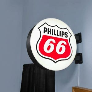 Puede incluir: Un letrero redondo de Phillips 66 con fondo blanco, borde negro y logotipo de escudo rojo y blanco. El letrero está montado en una pared con soportes negros y un cable de alimentación.