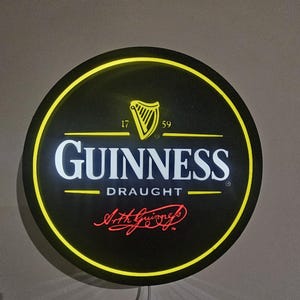 May include: Round black Guinness Draught sign with a yellow border and the Guinness logo. The logo includes a harp, the text "GUINNESS DRAUGHT", and the signature "Arth Guinness". The sign has a yellow illuminated border.
