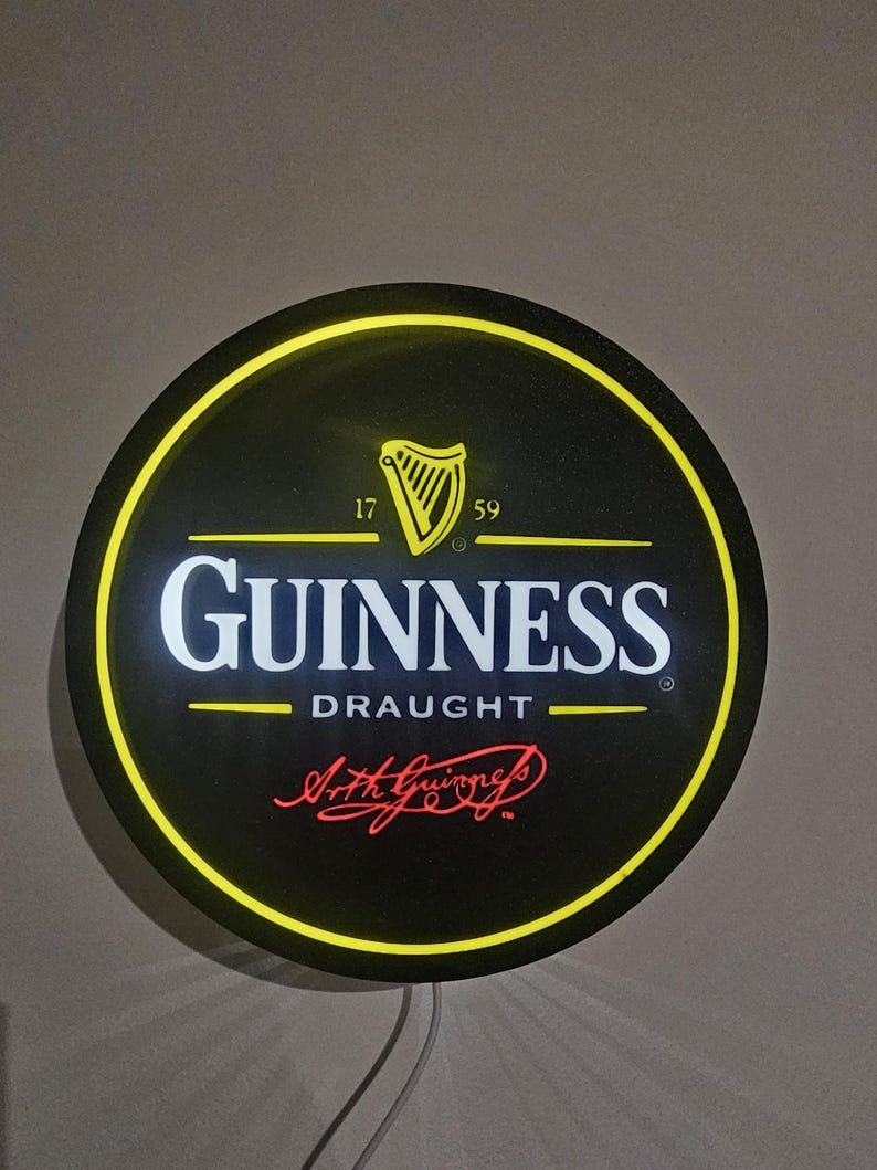 May include: Round black Guinness Draught sign with a yellow border and the Guinness logo. The logo includes a harp, the text "GUINNESS DRAUGHT", and the signature "Arth Guinness". The sign has a yellow illuminated border.
