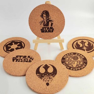 Peut inclure: Ensemble de dessous de verre ronds en liège avec des motifs Star Wars. Les dessous de verre présentent des logos et des illustrations de personnages, dont Dark Vador et l'Étoile de la Mort. Le texte "STAR WARS" est visible sur certains dessous de verre.