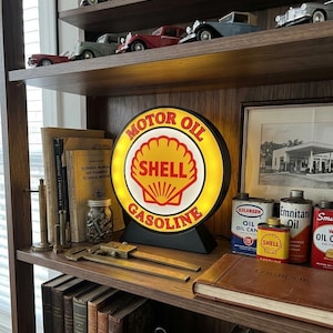 May include: A display of vintage items, including a Shell Motor Oil sign with the words "MOTOR OIL" and "GASOLINE". The illuminated sign is on a shelf with model cars, books, and vintage oil cans. The color scheme includes yellow, red, and black.
