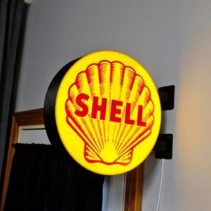 Peut inclure: Un panneau Shell rond et lumineux, avec un fond jaune et un motif de coquillage rouge. Le mot "SHELL" est affiché en rouge. Le panneau est monté sur un support noir.