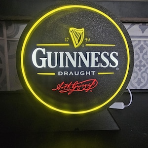 Peut inclure: Panneau Guinness Draught rond avec un fond noir et une bordure jaune n&eacute;on. Le logo Guinness est en blanc, avec une harpe dor&eacute;e et le texte "Arth Guinness" en rouge. Le panneau est &eacute;clair&eacute; d'une lueur jaune.
