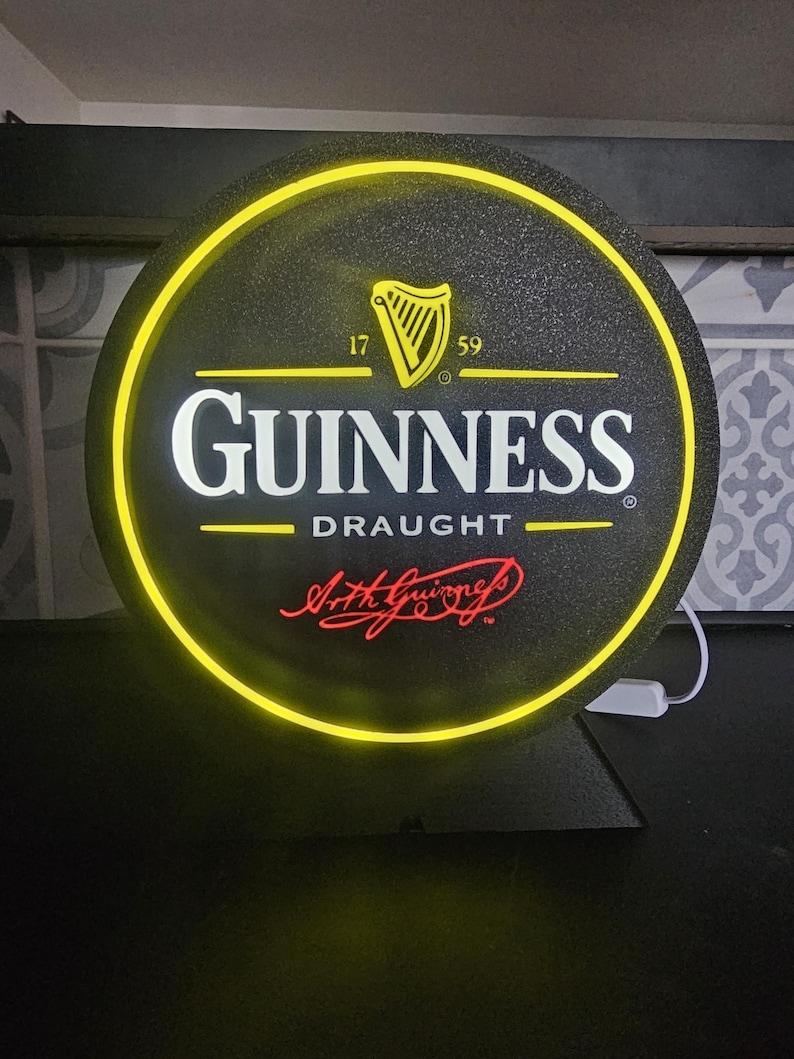 Peut inclure: Panneau Guinness Draught rond avec un fond noir et une bordure jaune n&eacute;on. Le logo Guinness est en blanc, avec une harpe dor&eacute;e et le texte "Arth Guinness" en rouge. Le panneau est &eacute;clair&eacute; d'une lueur jaune.