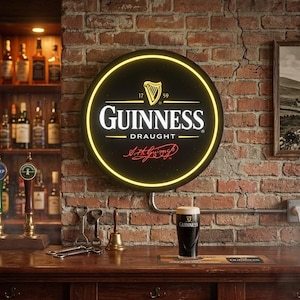 May include: A round Guinness Draught sign with a yellow neon border is mounted on a brick wall. The sign features the Guinness logo and a harp symbol. A pint of Guinness sits on a wooden bar, with bottles and taps in the background.