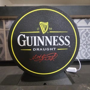Peut inclure: Panneau rond Guinness Draught avec fond noir et bordure jaune. Le logo Guinness est en blanc, avec un symbole de harpe et le texte "17 59". Le panneau est mont&eacute; sur un socle noir.