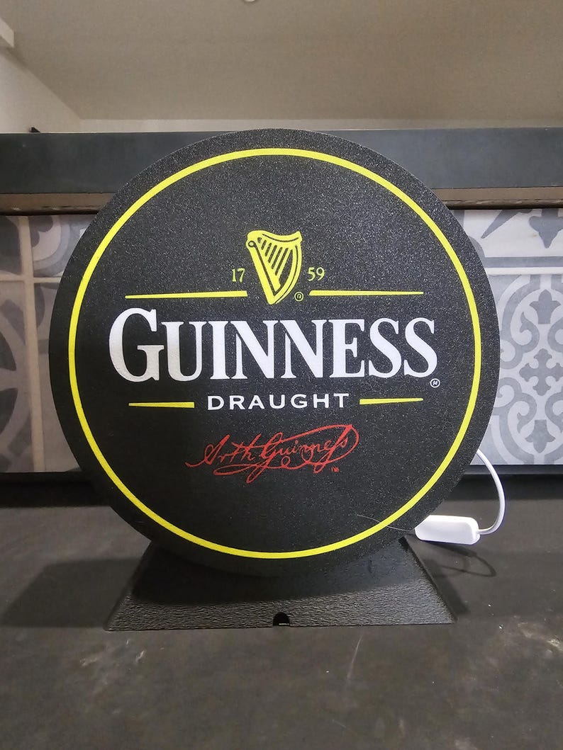 Peut inclure: Panneau rond Guinness Draught avec fond noir et bordure jaune. Le logo Guinness est en blanc, avec un symbole de harpe et le texte "17 59". Le panneau est mont&eacute; sur un socle noir.