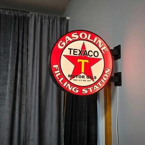 Puede incluir: Un letrero de gasolinera Texaco Gasoline vintage. El letrero redondo presenta un borde rojo con texto blanco y una estrella roja central con una "T" amarilla. El letrero está montado sobre un soporte negro.